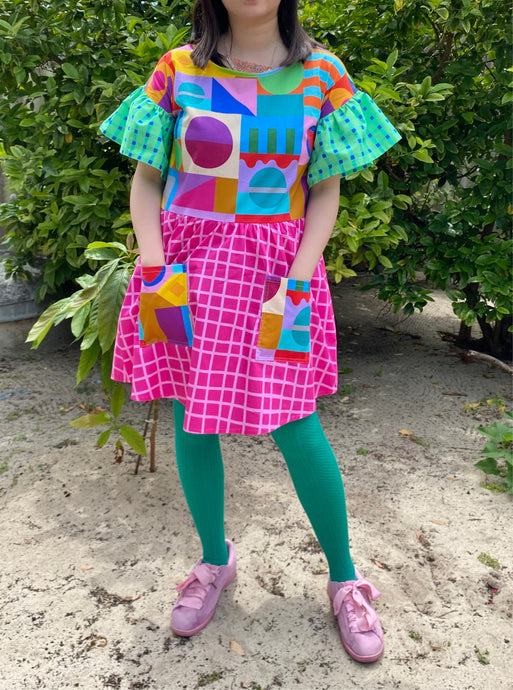 Dress with multicoloured geometric print bodice and pockets, frilly green and blue gingham sleeves and hot pink check skirt. Worn with green tights.