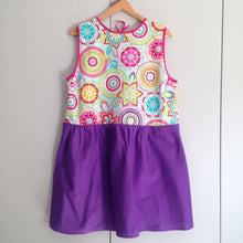 Load image into Gallery viewer, Front view of dress on a wooden hanger. The dress has a purple gathered skirt and sleeveless multicoloured bodice. The bodice has a geometric pattern that looks floral.

