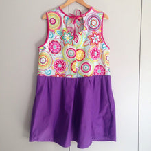 Load image into Gallery viewer, Back view of dress on a wooden hanger. The dress has a purple gathered skirt and sleeveless multicoloured bodice. The bodice has a geometric pattern that looks floral. The back of the neck has a keyhole and tie.
