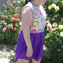 Load image into Gallery viewer, Shot of dress on a model walking through a rose garden. The dress has a purple gathered skirt and sleeveless multicoloured bodice. The bodice has a geometric pattern that looks floral.
