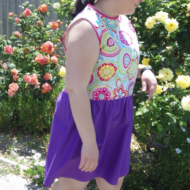 Shot of dress on a model walking through a rose garden. The dress has a purple gathered skirt and sleeveless multicoloured bodice. The bodice has a geometric pattern that looks floral.