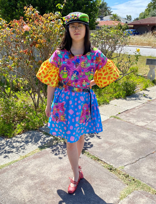 Dress with pink mermaid print bodice and pockets, yellow and orange sleeves and blue and pink skirt. Worn with denim rainbow belt.